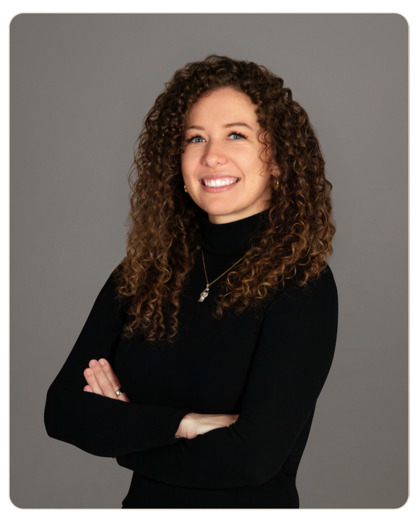 Smiling woman with curly brown hair in a black turtleneck, arms crossed against a gray studio background.
