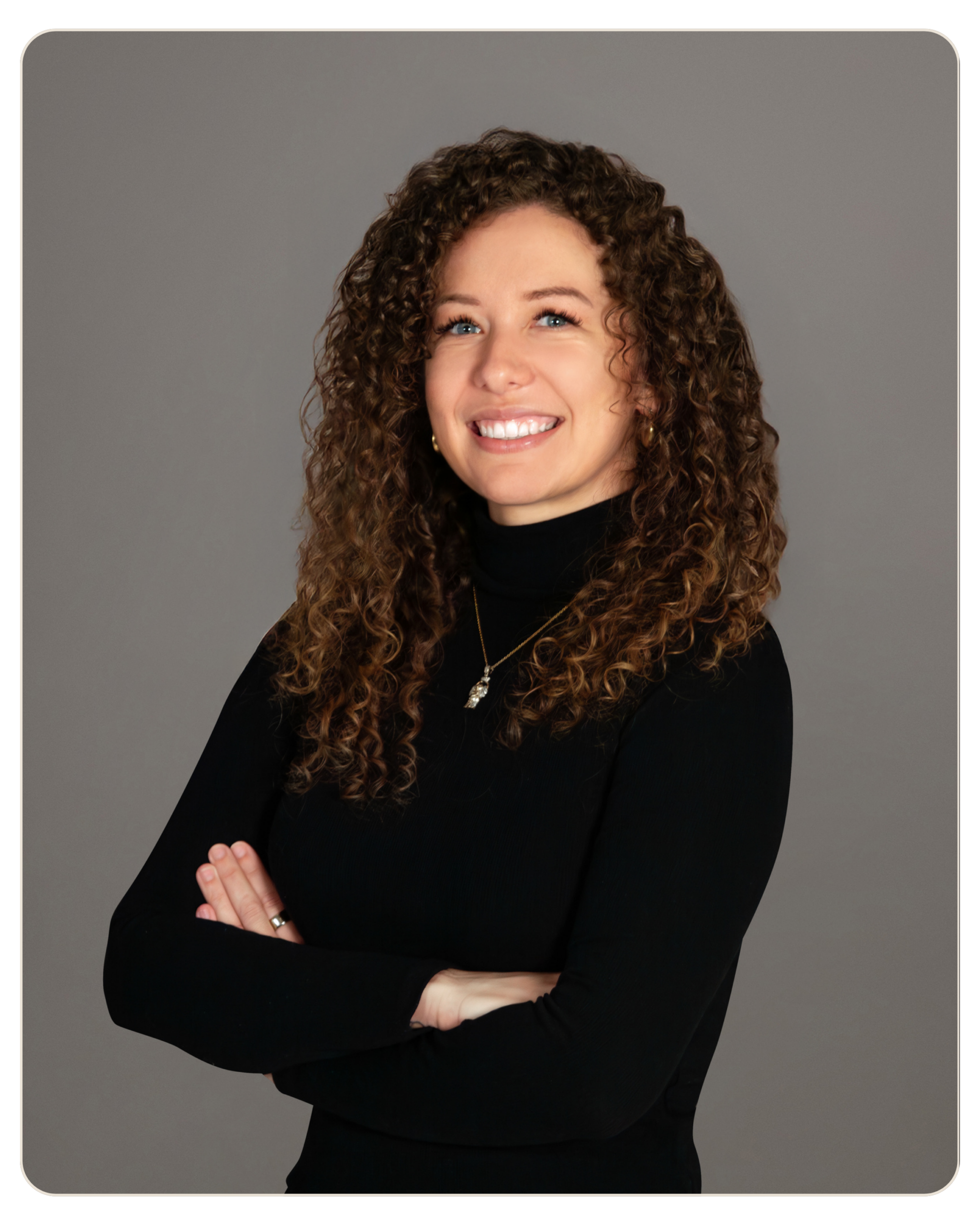 Smiling woman with curly brown hair in a black turtleneck, arms crossed against a gray studio background.