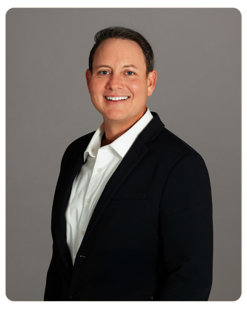Professional headshot of a smiling man in a dark blazer and white shirt against a gray studio backdrop.