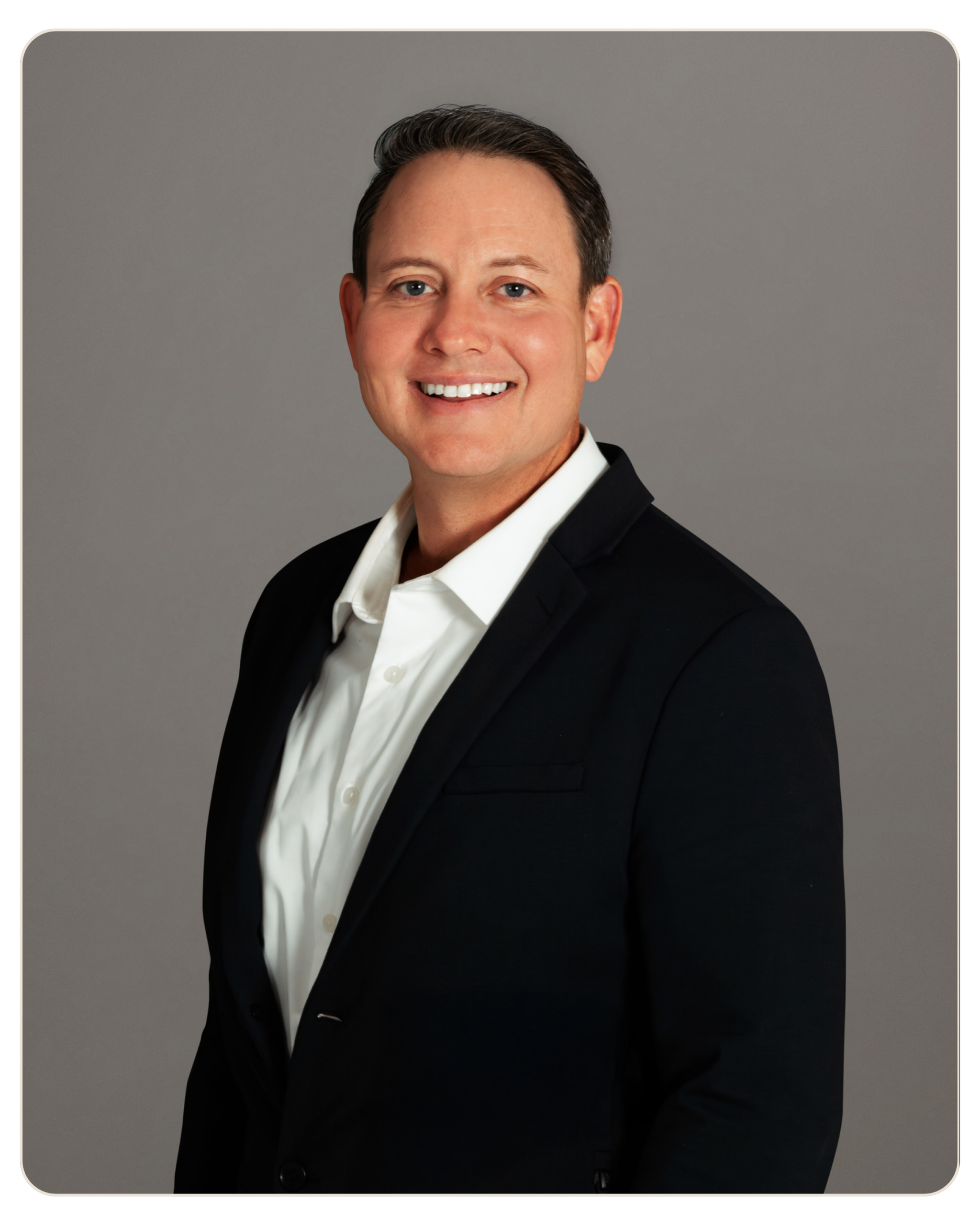 Professional headshot of a smiling man in a dark blazer and white shirt against a gray studio backdrop.