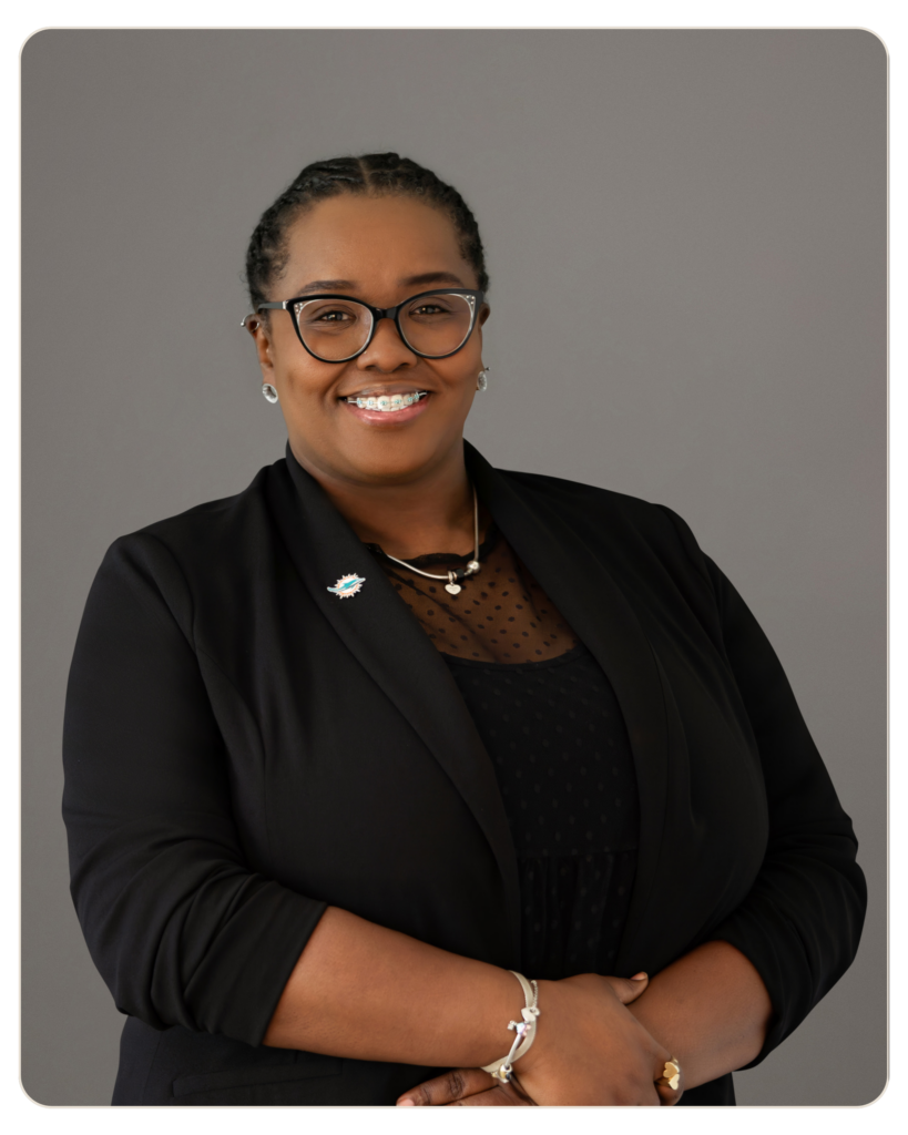 Beacon Recovery - Brittany Tee Owens Smiling Black woman in a black blazer with glasses, arms crossed, neutral gray background.