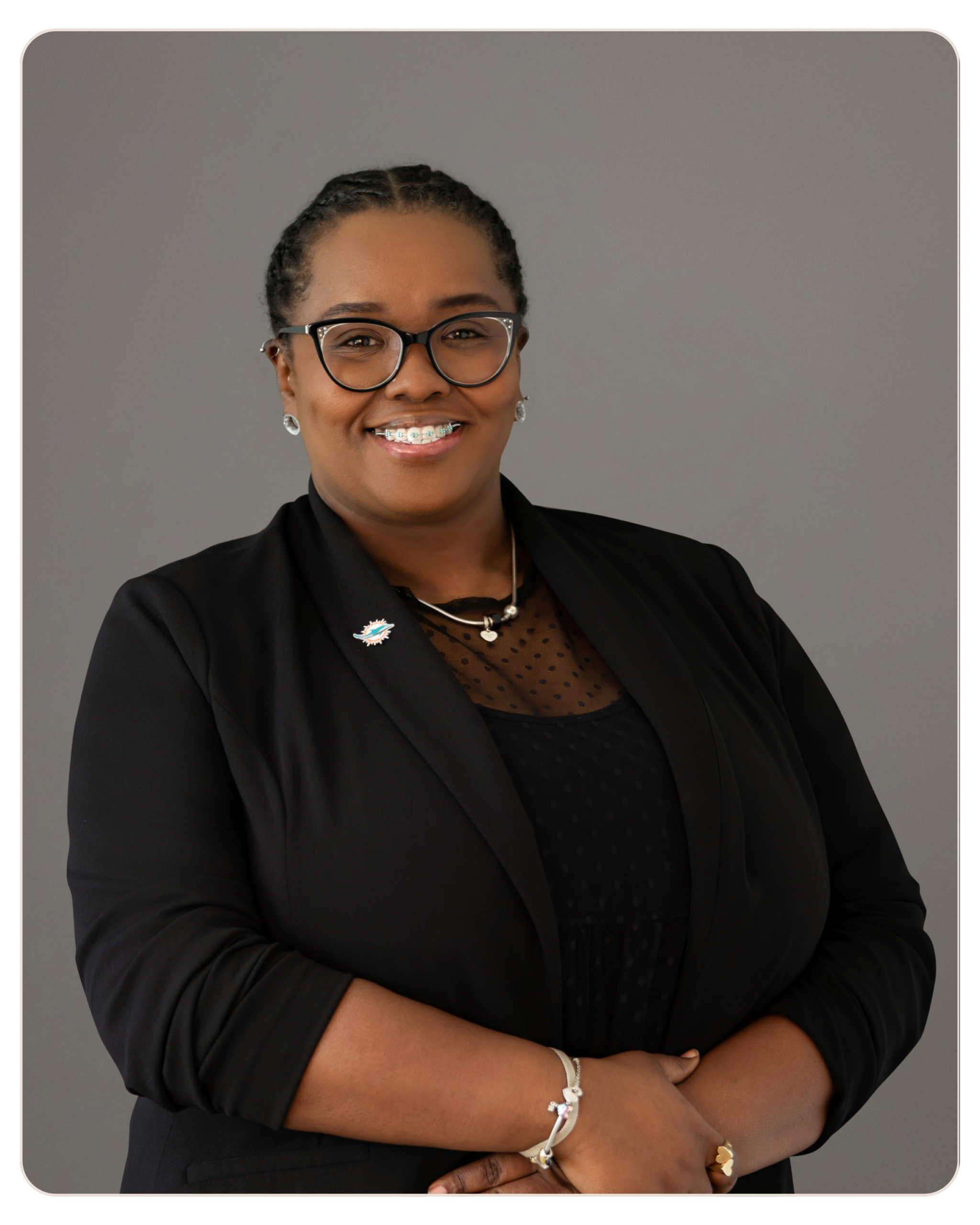 Beacon Recovery - Brittany Tee Owens Smiling Black woman in a black blazer with glasses, arms crossed, neutral gray background.