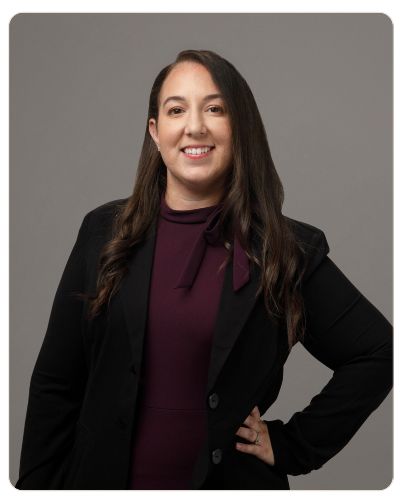 Flyland Crystal Donato Professional woman in a black blazer and burgundy top, standing with hand on hip against a gray studio background, smiling.
