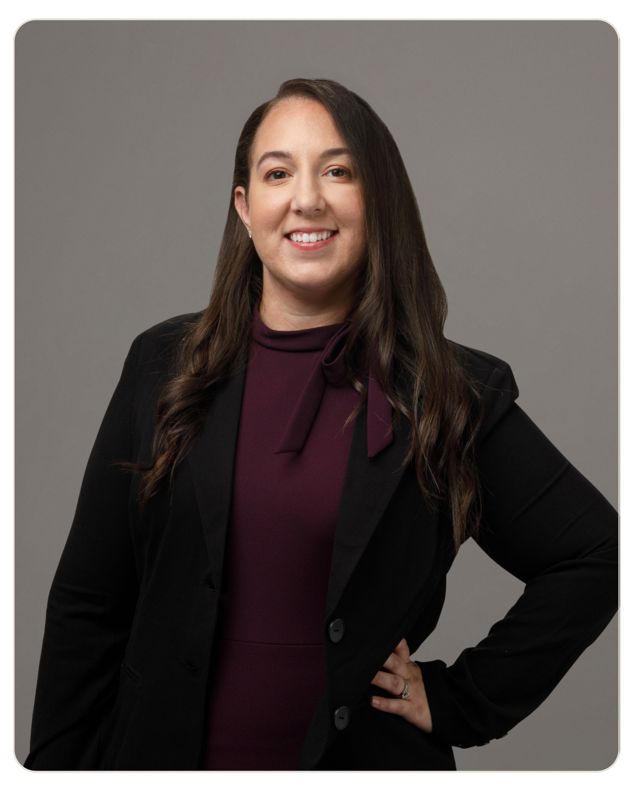 Flyland Crystal Donato Professional woman in a black blazer and burgundy top, standing with hand on hip against a gray studio background, smiling.