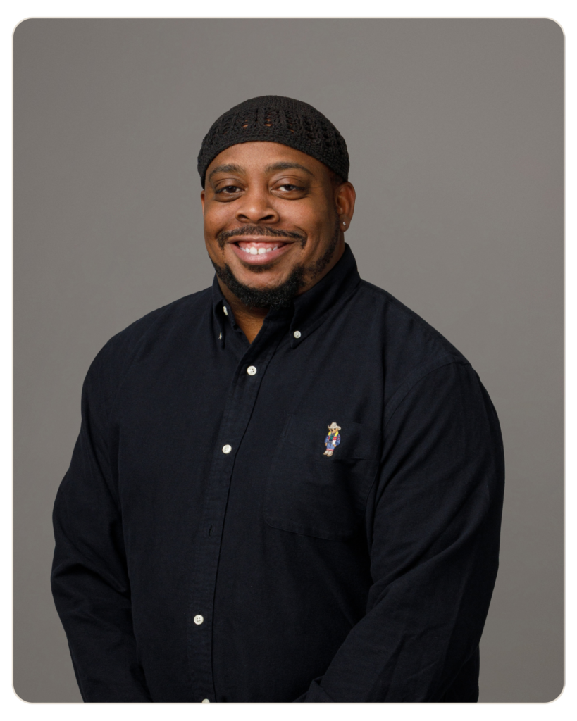 Smiling man wearing a black knit cap and dark button-down shirt against a gray studio background.