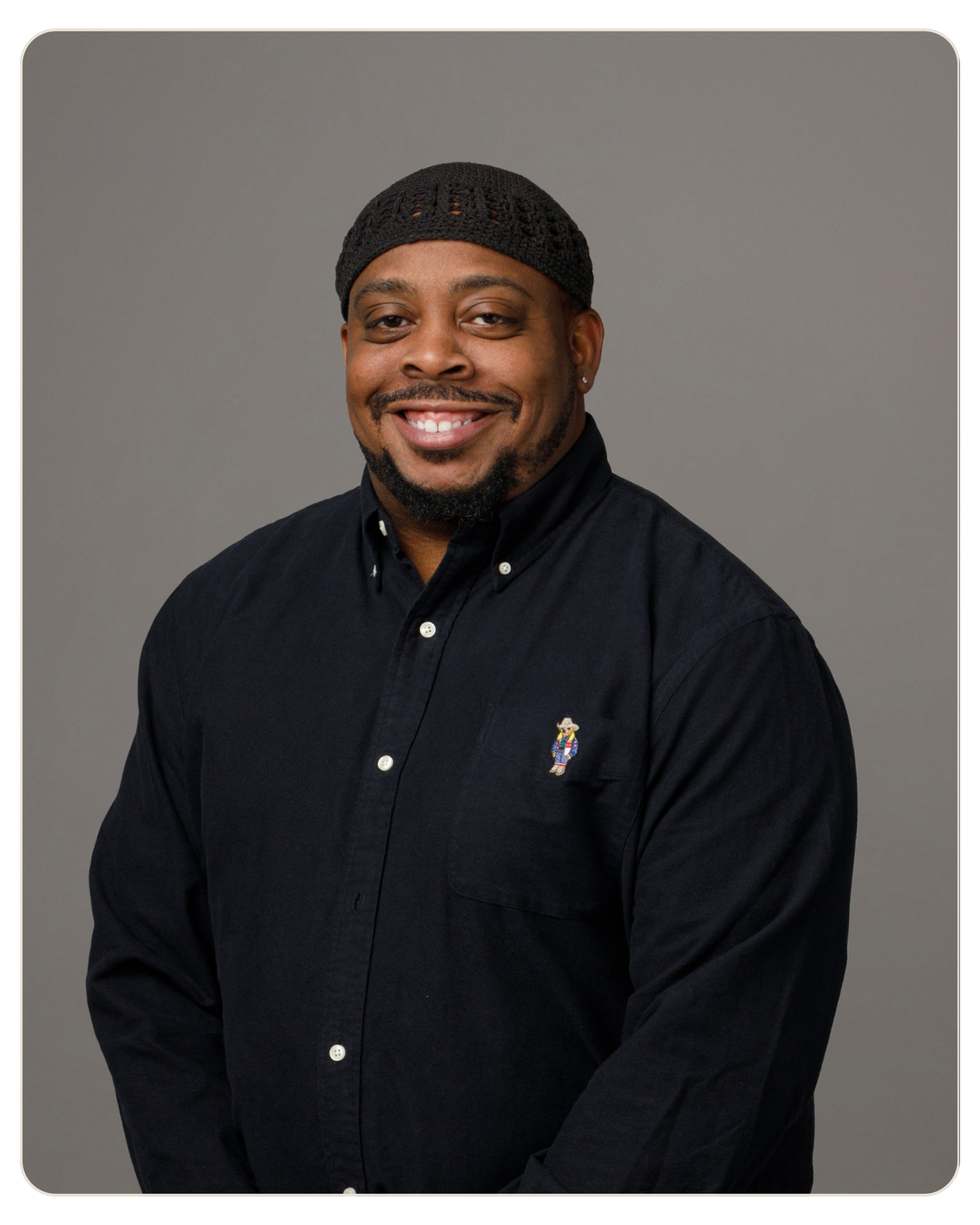 Smiling man wearing a black knit cap and dark button-down shirt against a gray studio background.