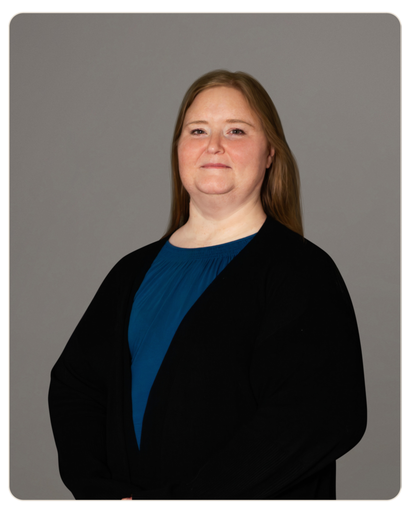 Jodie Simpson, LMFT Portrait of a smiling woman with light brown hair wearing a blue top and black blazer against a gray backdrop, arms crossed lightly to the side.