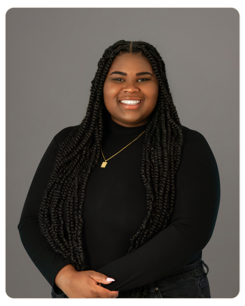 Smiling woman with long braided hair, wearing a black top and gold necklace, against a gray backdrop. Smiling woman with long braids, wearing a black top and gold necklace against a gray studio background.