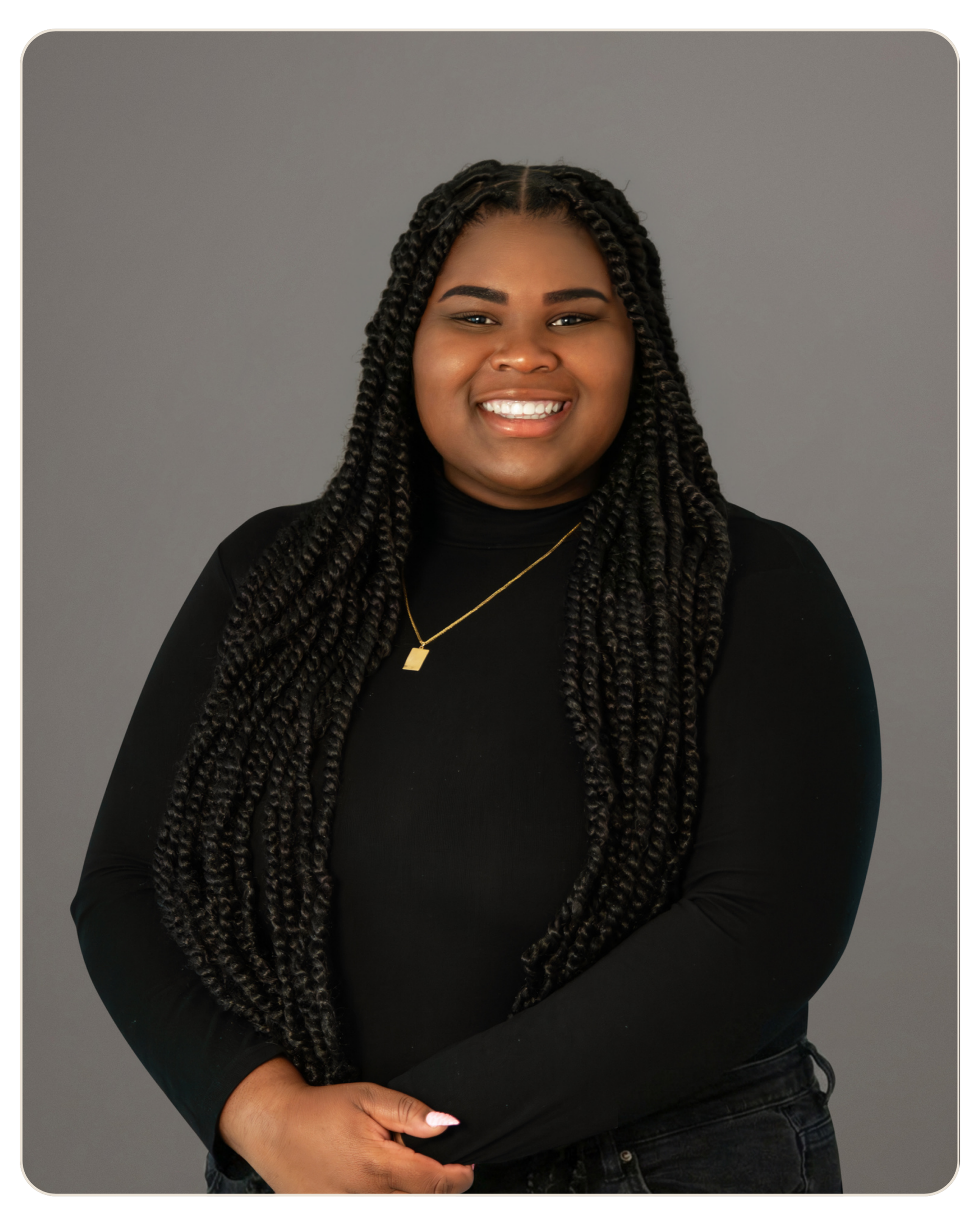 Smiling woman with long braided hair, wearing a black top and gold necklace, against a gray backdrop. Smiling woman with long braids, wearing a black top and gold necklace against a gray studio background.