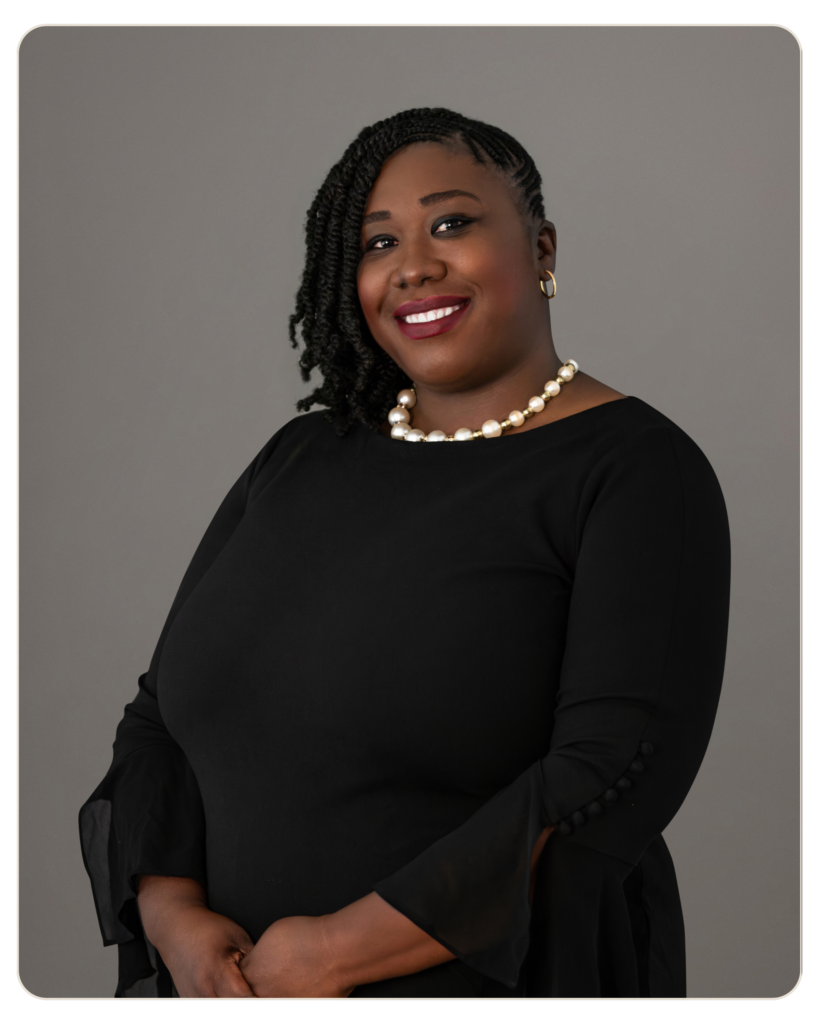 Beacon Recovery | Keshia Frederique, MSW Portrait of a smiling Black woman with braided hair, wearing a black dress and a pearl necklace against a gray background, studio lighting.