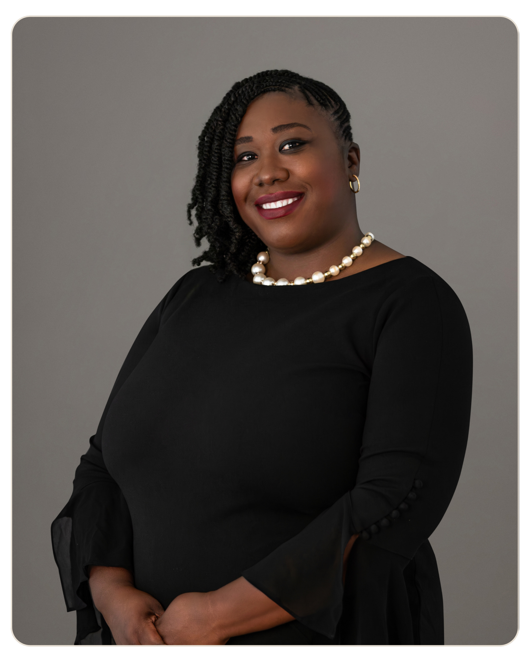 Beacon Recovery | Keshia Frederique, MSW Portrait of a smiling Black woman with braided hair, wearing a black dress and a pearl necklace against a gray background, studio lighting.