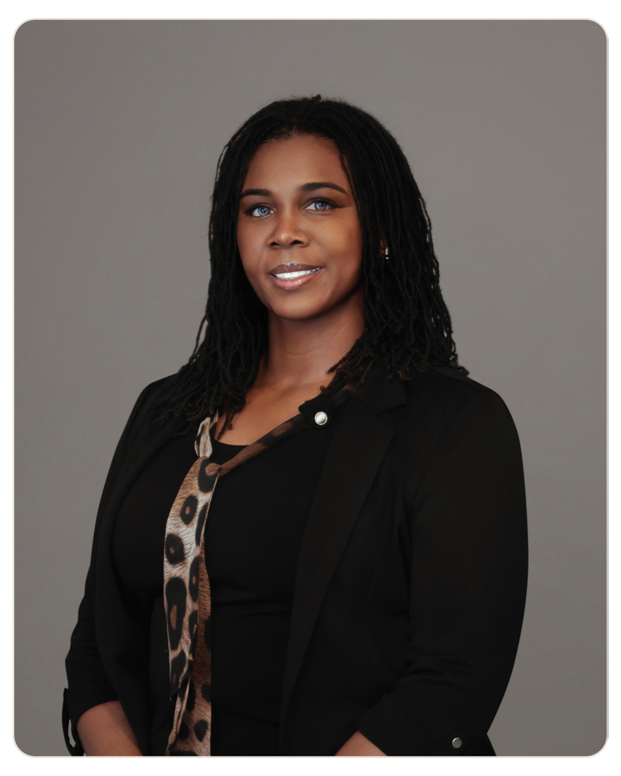 Kizzy Thomas, MSW Primary Therapist Head-and-shoulders portrait of a smiling African American woman with braided hair, wearing a black blazer and leopard scarf, gray background.