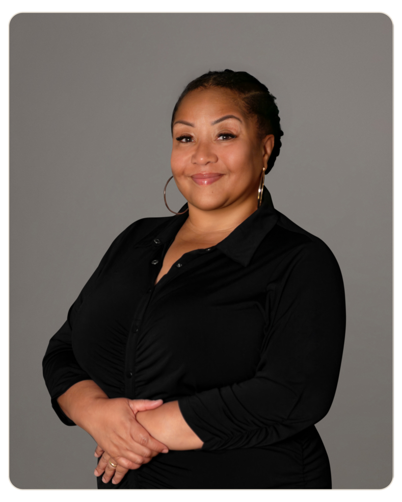 Magaly Padilla, Primary Therapist at Beacon Smiling woman with braided hair, wearing a black blouse and gold hoop earrings, arms crossed in a studio portrait against a gray background.