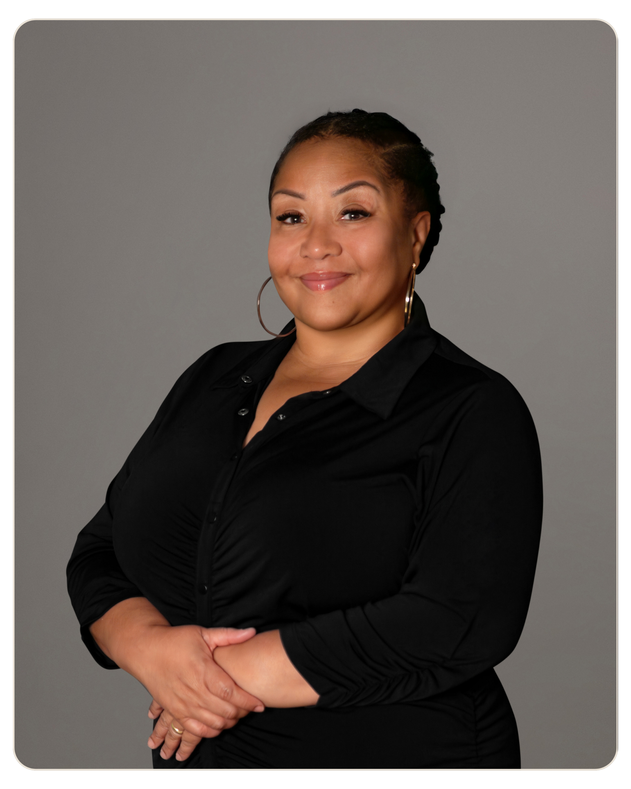 Magaly Padilla, Primary Therapist at Beacon Smiling woman with braided hair, wearing a black blouse and gold hoop earrings, arms crossed in a studio portrait against a gray background.