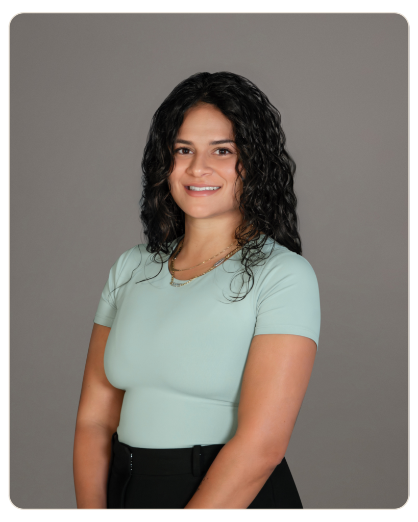Rafaela Buenaventura Executive Director of Programming Portrait of a smiling woman with dark, curly hair wearing a light green top and black pants, posed against a gray studio backdrop.