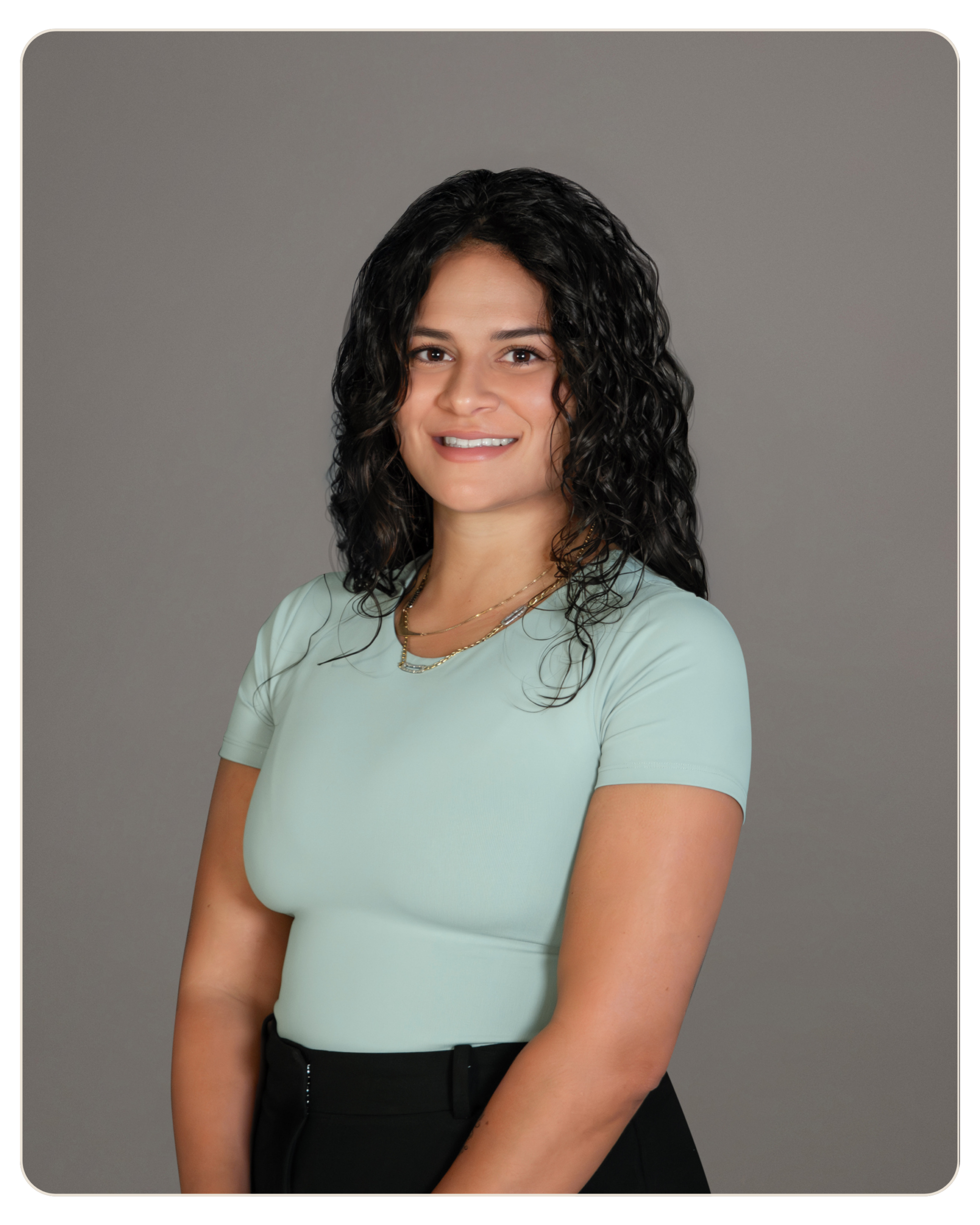Rafaela Buenaventura Executive Director of Programming Portrait of a smiling woman with dark, curly hair wearing a light green top and black pants, posed against a gray studio backdrop.