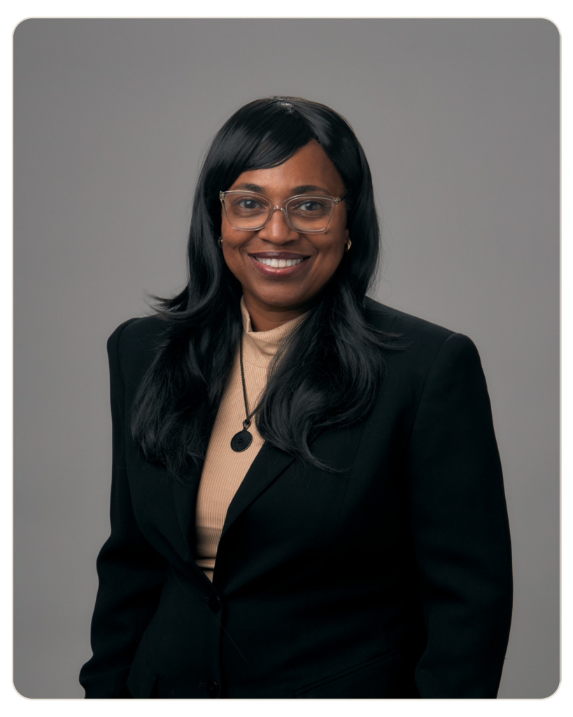 Robin Jeffrey Recovery Coach Head-and-shoulders portrait of a smiling Black woman with long black hair, wearing glasses, a beige sweater, and a black blazer against a gray background.