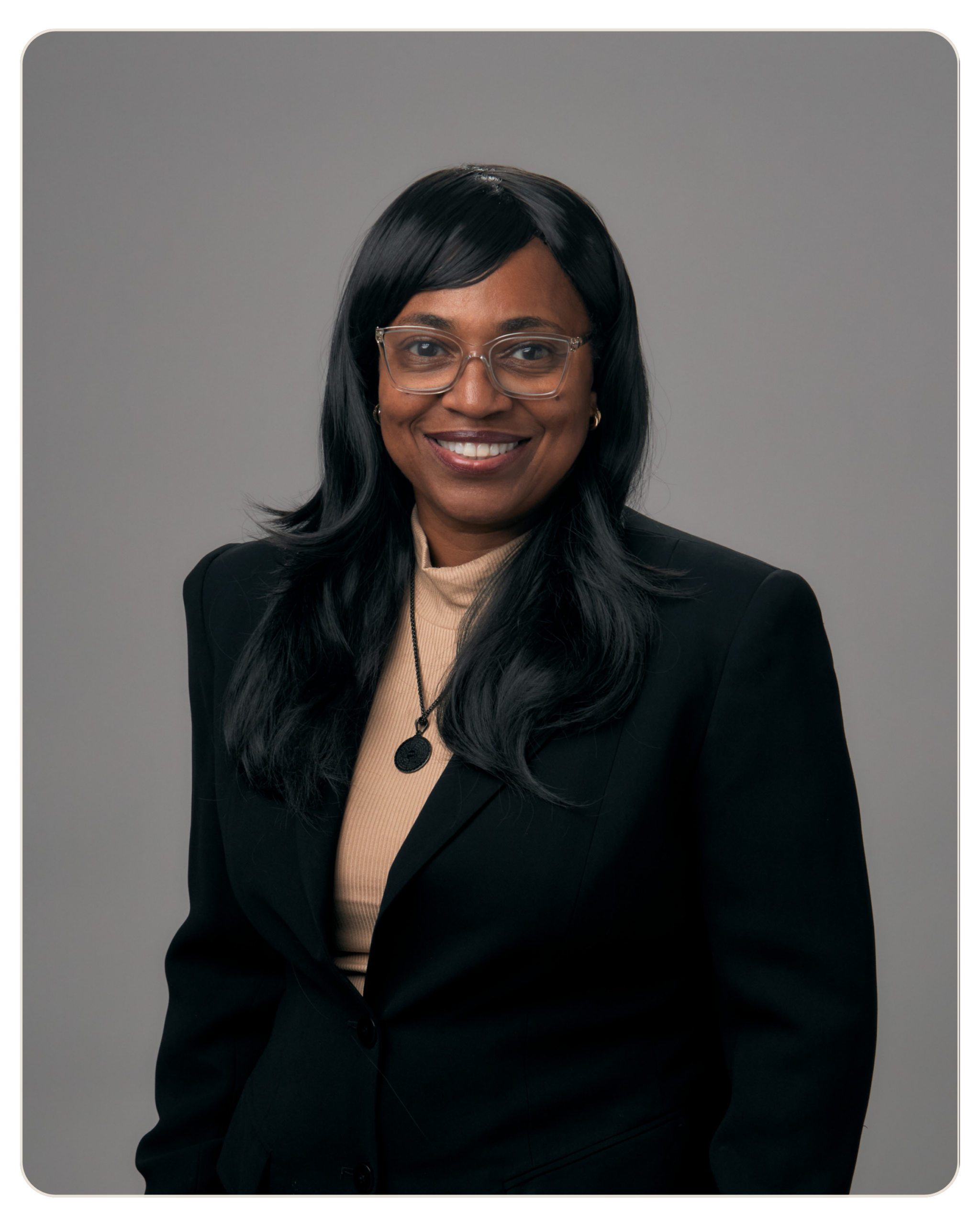 Robin Jeffrey Recovery Coach Head-and-shoulders portrait of a smiling Black woman with long black hair, wearing glasses, a beige sweater, and a black blazer against a gray background.