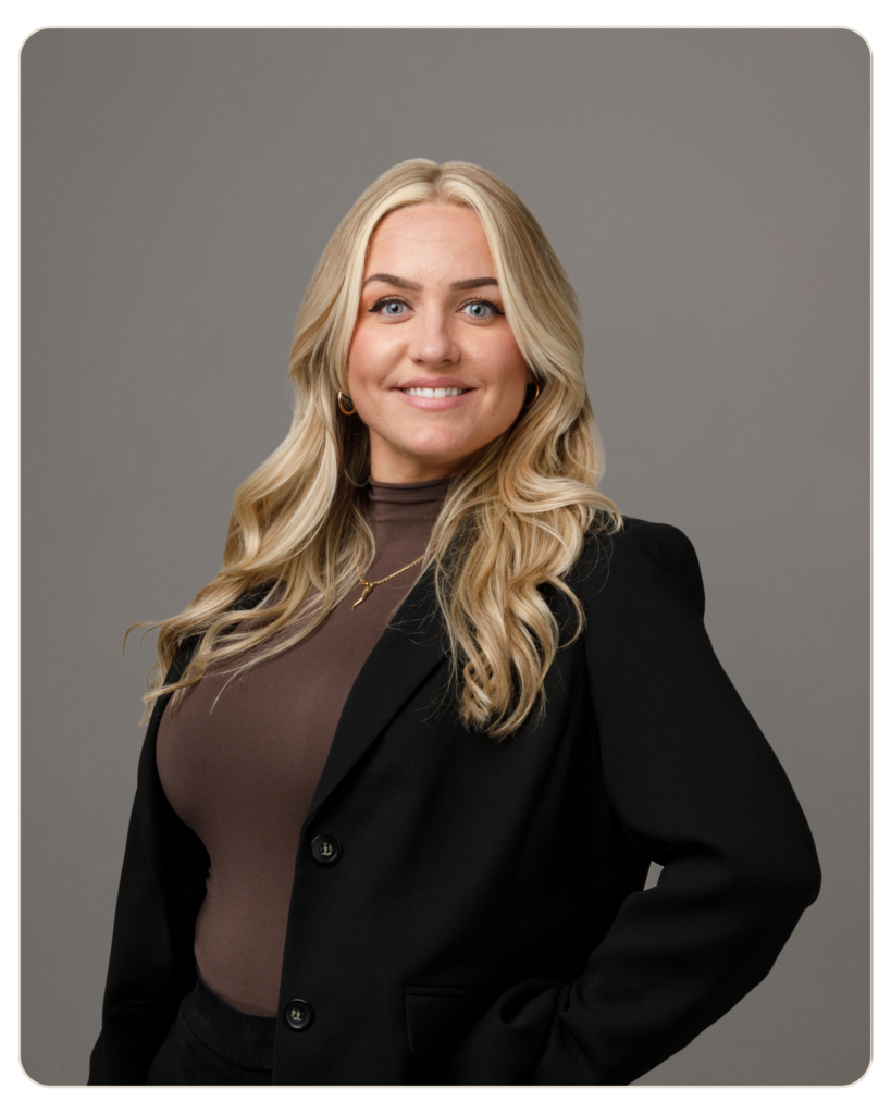 Samantha Felice Director of HR Portrait of a smiling woman with long blonde hair, wearing a black blazer over a brown top, against a gray studio background.