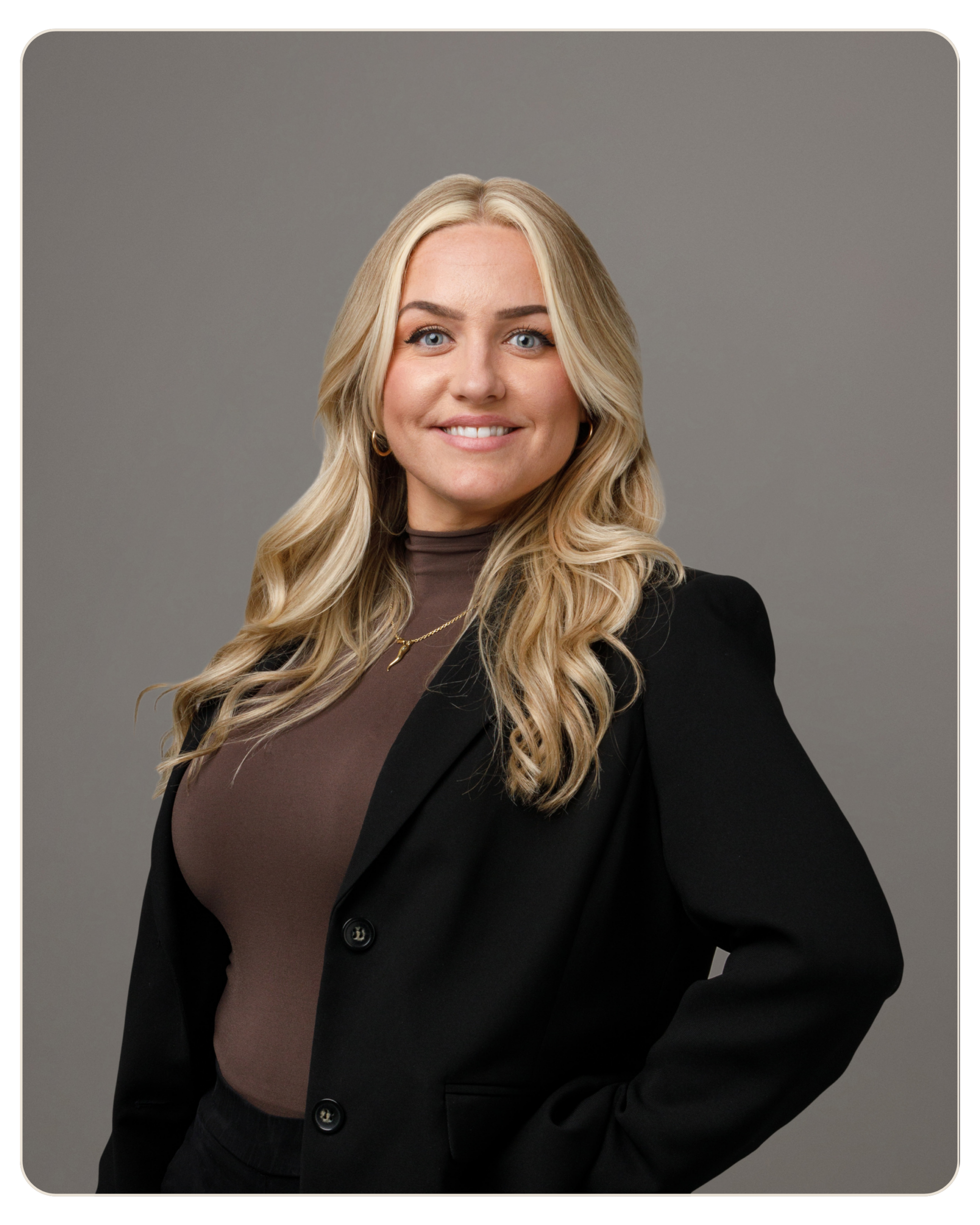 Samantha Felice Director of HR Portrait of a smiling woman with long blonde hair, wearing a black blazer over a brown top, against a gray studio background.