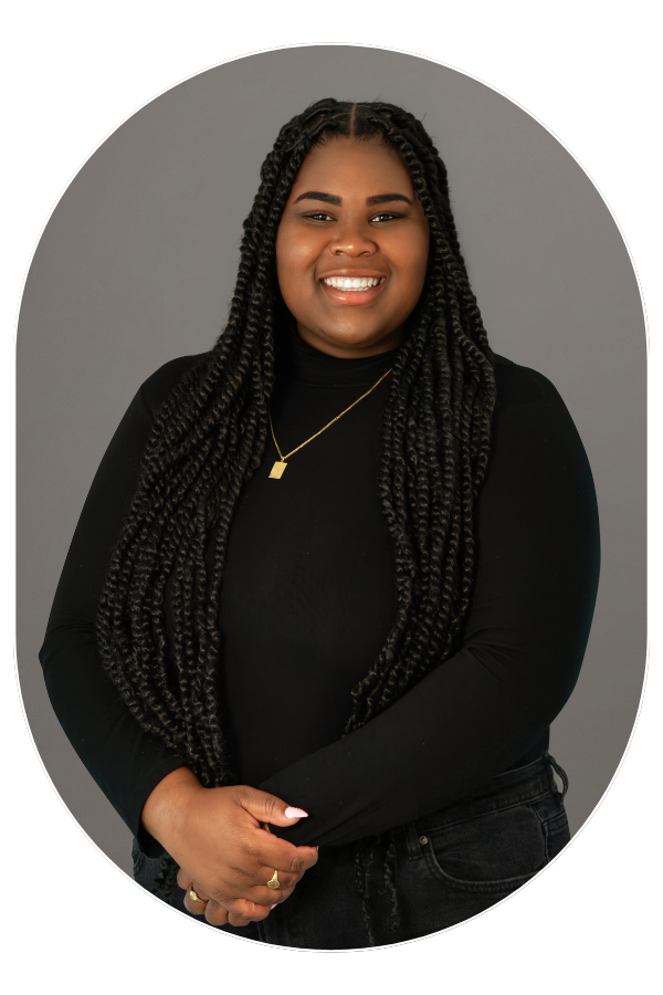 Smiling woman with long braided hair, wearing a black top and gold necklace, against a gray backdrop.