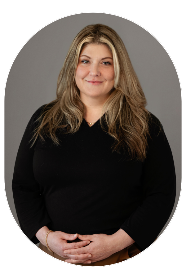 Portrait of a smiling woman with long blonde hair wearing a black top, hands gently clasped in front against a gray backdrop.